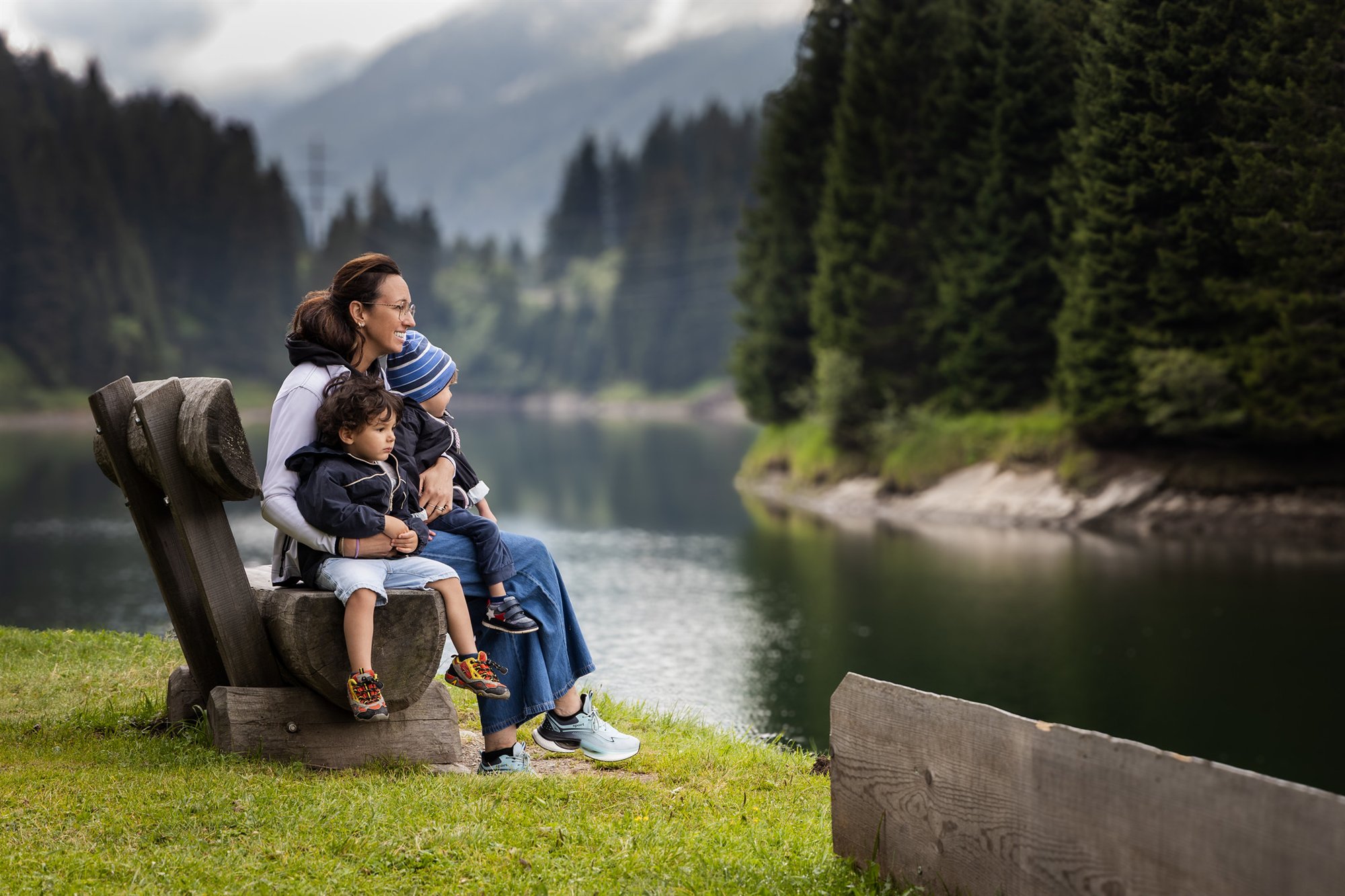 Una donna seduta su una panchina di legno con due bambini davanti a un lago di montagna